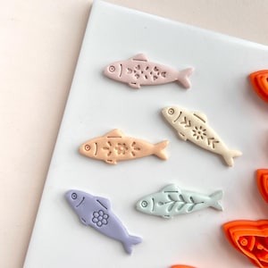 Peut inclure: Collection de découpoirs à biscuits en forme de poisson aux couleurs pastel. Les découpoirs sont dans les tons de rose, pêche, crème, lavande et vert menthe. Chaque poisson a un motif floral ou de feuille unique. Des découpoirs en forme de poisson orange sont également visibles.
