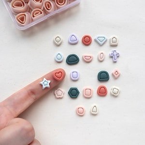 May include: A collection of small, multi-coloured geometric clay cutters in a clear plastic storage container, accompanied by various finished clay shapes in shades of blue, green, pink, and white, including stars, hearts, and crosses.