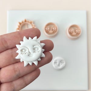 Op de afbeelding: Een witte klei zon met een lachend gezicht en stekelige stralen, vastgehouden in een hand. Andere kleistukken zijn een zonvormige uitsteker en maan- en zongezichtsontwerpen. De items liggen op een wit oppervlak.