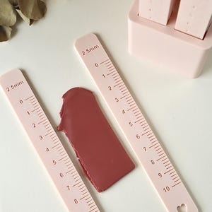 Puede incluir: Primer plano de una regla rosa pálido con medidas en milímetros, junto a un objeto rectangular rojo oscuro. La regla tiene un recorte en forma de corazón en el extremo. La regla mide hasta 10 mm.