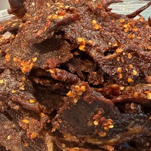 Puede incluir: Primer plano de un mont&oacute;n de cecina de res de color marr&oacute;n oscuro con hojuelas de pimiento rojo. La cecina est&aacute; en un recipiente de pl&aacute;stico transparente.