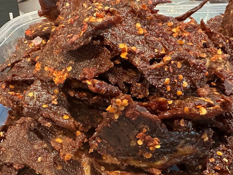 Puede incluir: Primer plano de un mont&oacute;n de cecina de res de color marr&oacute;n oscuro con hojuelas de pimiento rojo. La cecina est&aacute; en un recipiente de pl&aacute;stico transparente.
