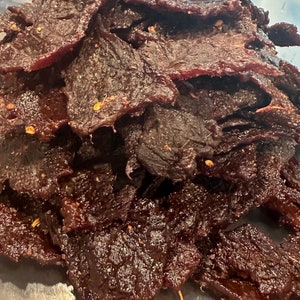 Puede incluir: Primer plano de un recipiente lleno de cecina de res de color marr&oacute;n oscuro. La cecina est&aacute; dispuesta en una pila y parece estar sazonada con especias.