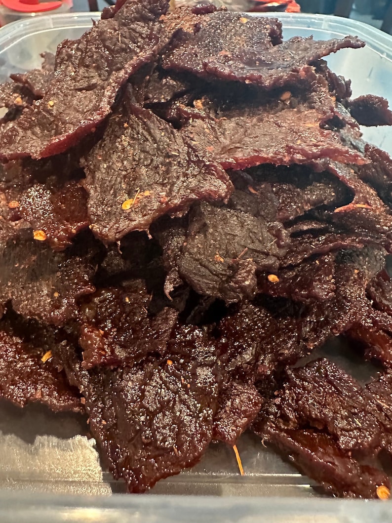 Puede incluir: Primer plano de un recipiente lleno de cecina de res de color marr&oacute;n oscuro. La cecina est&aacute; dispuesta en una pila y parece estar sazonada con especias.