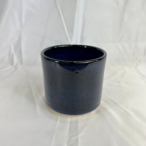 May include: A dark blue ceramic planter with a smooth, glossy finish. The planter is cylindrical in shape and has a slightly rounded bottom.