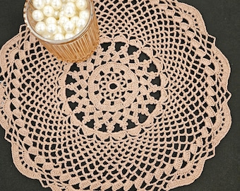 May include: A beige, crocheted doily with a scalloped edge. The doily has a circular design with an open, lacy pattern. A small, clear glass container filled with white pearls sits on the doily.