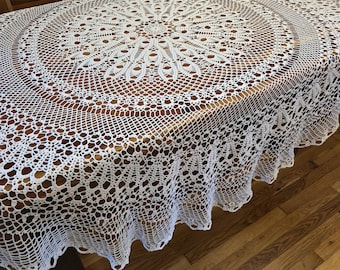 May include: A white, crocheted tablecloth with an elaborate circular pattern. The design includes a central motif and concentric rings of openwork, with a scalloped edge. The tablecloth is displayed on a wooden table, highlighting its delicate texture and craftsmanship.