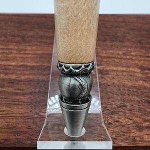 May include: Close-up of a wooden pen with a silver baseball-shaped cap and a decorative band. The pen is displayed on a clear acrylic stand.