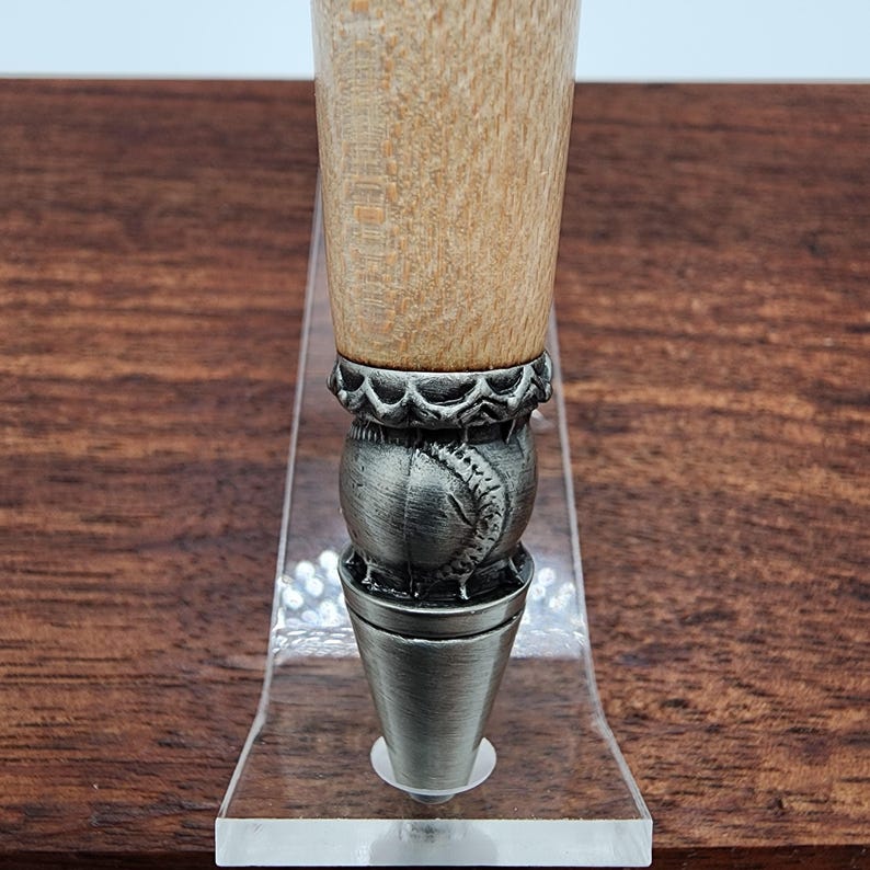 May include: Close-up of a wooden pen with a silver baseball-shaped cap and a decorative band. The pen is displayed on a clear acrylic stand.