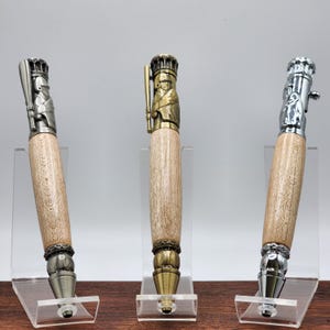 May include: Three wooden pens with metal baseball player toppers. The pens are displayed on clear acrylic stands. The toppers are in silver, gold, and brass.