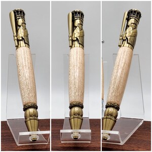 May include: A wooden pen with a gold-toned baseball player topper. The pen is displayed on a clear acrylic stand. The pen has a baseball-themed design at the bottom.