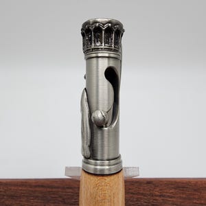 May include: A silver pen with a detailed, ornate crown-shaped top and a wooden barrel. The pen has a unique design with a small ball on the side.