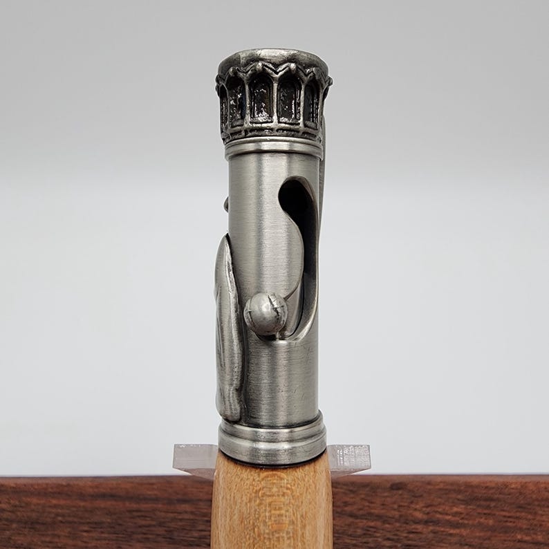 May include: A silver pen with a detailed, ornate crown-shaped top and a wooden barrel. The pen has a unique design with a small ball on the side.