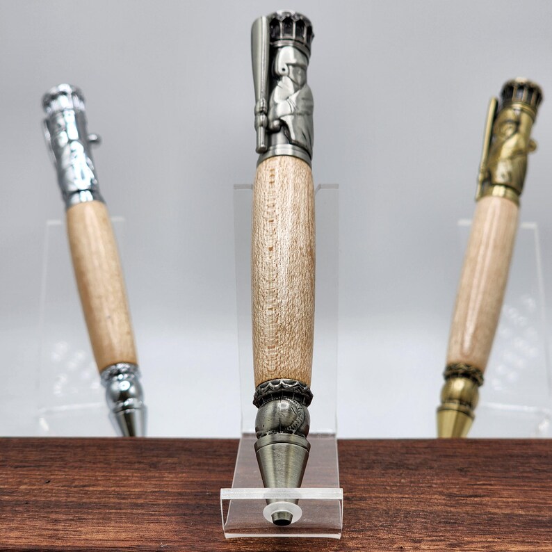 May include: Three wooden pens with metal baseball player toppers. The pens are on a wooden surface and are displayed on clear acrylic stands. The baseball player toppers are in different poses.