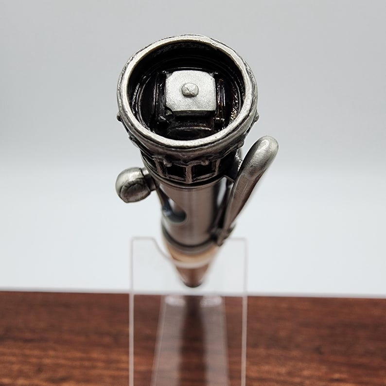 May include: A silver pen with a detailed design resembling a building. The pen has a black clip and a silver cap with a small, square button on top.