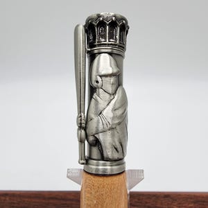 May include: A silver pen with a detailed baseball player design on the barrel. The pen has a wooden base.