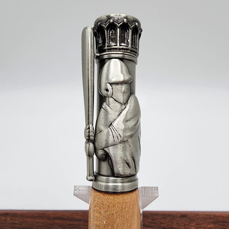 May include: A silver pen with a detailed baseball player design on the barrel. The pen has a wooden base.