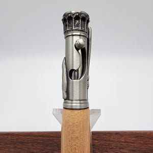 May include: A silver pen with a detailed design on the top and a wooden barrel. The pen is on a clear acrylic stand.