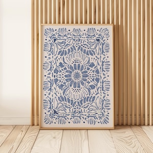 May include: A framed art print featuring a blue floral design on a cream background. The artwork showcases a central flower surrounded by a rectangular border of stylized flowers and leaves. The frame is a light wood color.