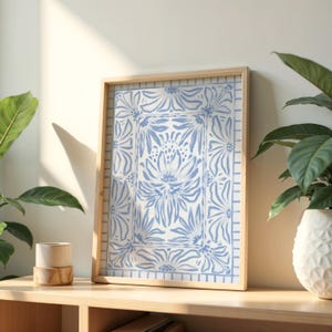 May include: A framed art print featuring a blue and white floral design. The artwork is in a light wooden frame and rests on a wooden shelf. A small ceramic cup and a potted plant are also on the shelf.