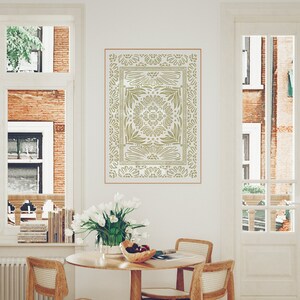May include: A stencil of a decorative design with a floral pattern in a light green color on a white wall. The stencil is framed by a thin brown line. The stencil is hanging above a wooden table with two chairs.
