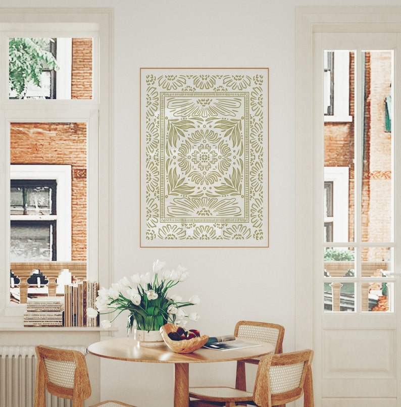 May include: A stencil of a decorative design with a floral pattern in a light green color on a white wall. The stencil is framed by a thin brown line. The stencil is hanging above a wooden table with two chairs.