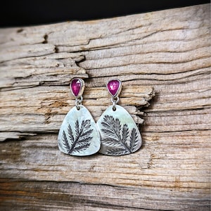 May include: A pair of silver earrings with a botanical design. Each earring features a teardrop-shaped pendant with a detailed fern-like pattern. The earrings are topped with a small, vibrant pink gemstone. The earrings are displayed on a wooden surface.