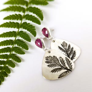 May include: Silver earrings with a teardrop-shaped ruby gemstone and a fern leaf design. The earrings have a post back and a dangling, engraved pendant. A green fern leaf is in the background.
