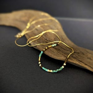 Peut inclure: Un collier délicat en chaîne dorée avec une petite rangée de perles turquoise, blanches et brunes. Le collier est présenté sur un morceau de bois flotté.