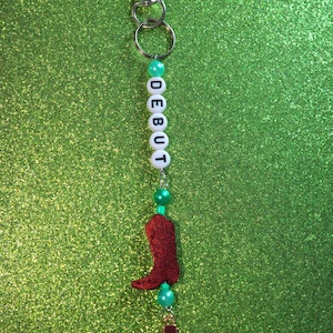 May include: A keychain with a silver clasp, white letter beads spelling "DEBUT", and green beads. Charms include a red cowboy boot and a turquoise ukulele. The background is a sparkly green.