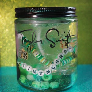 May include: A clear glass jar with a black lid filled with green liquid and decorative elements. Inside are beaded bracelets with the text "Taylor Swift" and "Tim McGraw". The jar is on a teal surface.