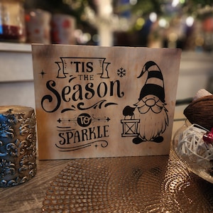 Może przedstawiać: Drewniany znak z rustykalnym brązowym tłem i czarnymi napisami. Znak głosi "'Tis the Season to Sparkle" i przedstawia kreskówkowego gnoma trzymającego latarnię.