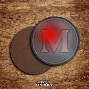 Könnte beinhalten: Ein grauer Untersetzer mit einem roten Leuchten hinter dem großen Buchstaben "M" und einem dunkelgrauen Rand. Ein zweiter, brauner Untersetzer liegt daneben. Der Text "dtox creative" befindet sich unten links.