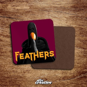 May include: Square coaster featuring a black bird-like figure with an orange beak and the word "FEATHERS" in yellow. The background is burgundy. A second, plain brown coaster is next to it. The image is on a wooden surface.