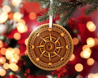 Puede incluir: Adorno navideño redondo y marrón con un diseño de rosa de los vientos. El adorno presenta una estrella central, fases lunares y bordes decorativos. Cuelga de una cinta blanca sobre un fondo borroso de luces y vegetación navideñas.