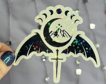 May include: A black and iridescent sticker with a crescent moon, a mountain range, and a sword. The moon is surrounded by a circle of stars and the wings of a bat. The sticker has a holographic finish.