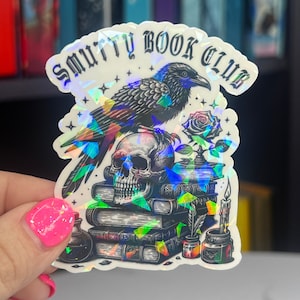 May include: A holographic sticker with a black raven perched on a skull, surrounded by a stack of books, a rose, and a candle. The sticker reads "Smutty Book Club".