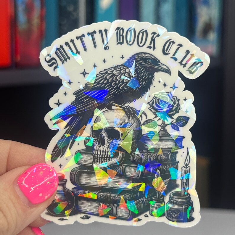 May include: A holographic sticker with a black raven perched on a skull and a stack of books. The sticker says "Smutty Book Club".