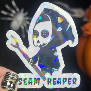 May include: A holographic sticker featuring a cartoon Grim Reaper holding a needle and thread. The sticker says "SEAM REAPER" in a holographic font.
