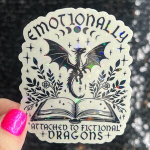 May include: A holographic sticker with a black and white illustration of a dragon emerging from an open book. The text reads "Emotionally Attached to Fictional Dragons".