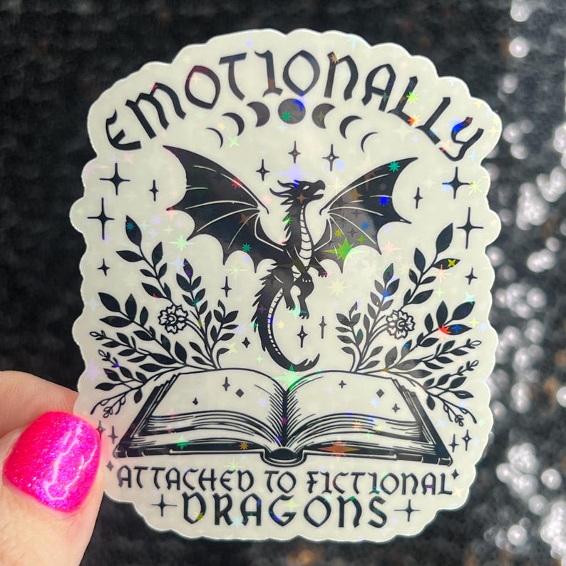 May include: A holographic sticker with a black and white illustration of a dragon emerging from an open book. The text reads "Emotionally Attached to Fictional Dragons".
