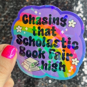 May include: A holographic sticker with a rainbow background and the text "Chasing that Scholastic Book Fair high". The sticker features a stack of books and a cloud shape.