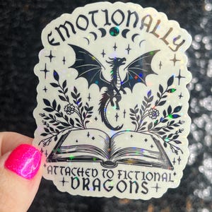 May include: A holographic sticker with a black and white illustration of a dragon, a book, and floral elements. The text reads "Emotionally Attached to Fictional Dragons".