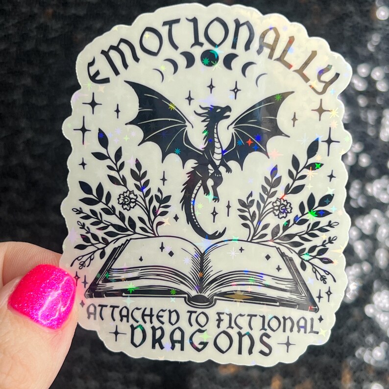 May include: A holographic sticker with a black and white illustration of a dragon, a book, and floral elements. The text reads "Emotionally Attached to Fictional Dragons".