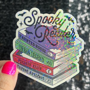Op de afbeelding: Een holografische sticker met de tekst "Spooky Reader" en een stapel boeken met de titels "Haunted House", "Urban Legend", "Psychotic Killer" en "Cursed Artifact".