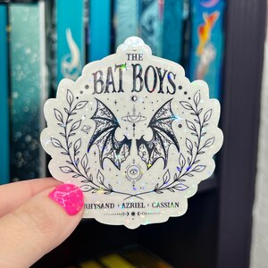 Pu&ograve; includere: Un adesivo bianco con finitura olografica che presenta un'illustrazione in bianco e nero di un paio di ali di pipistrello con una luna crescente e stelle. Il testo "THE BAT BOYS" &egrave; scritto in un font nero corsivo. Il testo "RHYSAND + AZRIEL + CASSIAN" &egrave; scritto in un font nero pi&ugrave; piccolo nella parte inferiore dell'adesivo.