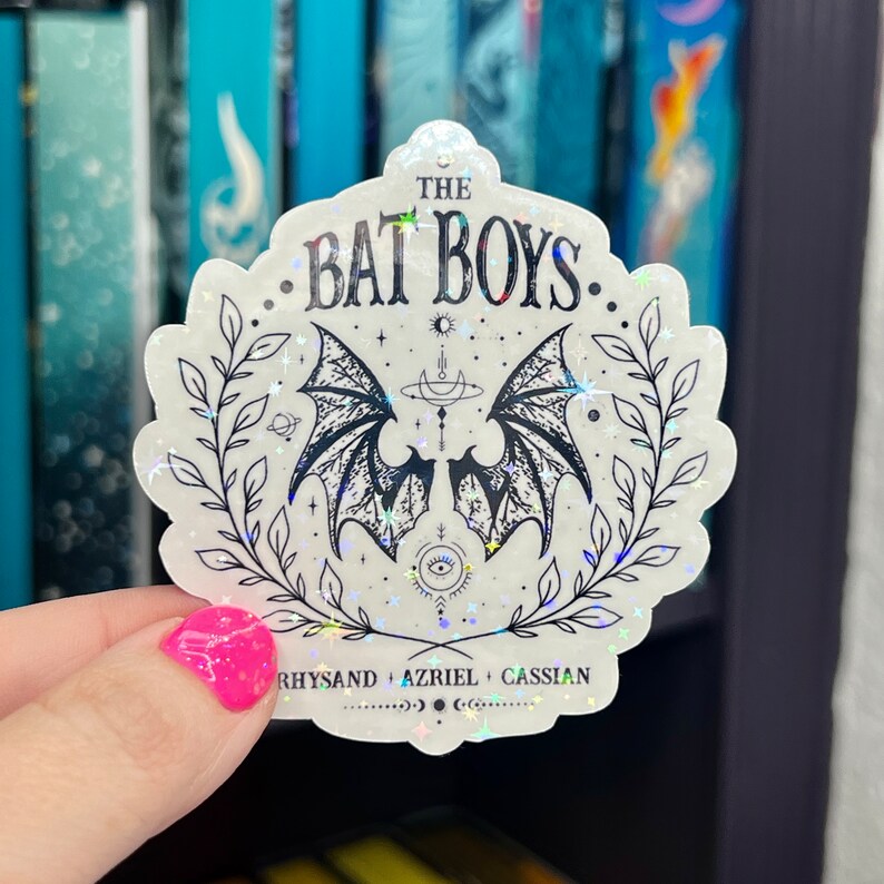 Pu&ograve; includere: Un adesivo bianco con finitura olografica che presenta un'illustrazione in bianco e nero di un paio di ali di pipistrello con una luna crescente e stelle. Il testo "THE BAT BOYS" &egrave; scritto in un font nero corsivo. Il testo "RHYSAND + AZRIEL + CASSIAN" &egrave; scritto in un font nero pi&ugrave; piccolo nella parte inferiore dell'adesivo.