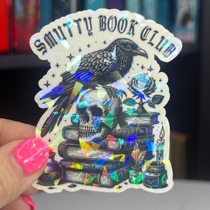 May include: A holographic sticker with a black raven, skull, and stack of books. The sticker says "Smutty Book Club".