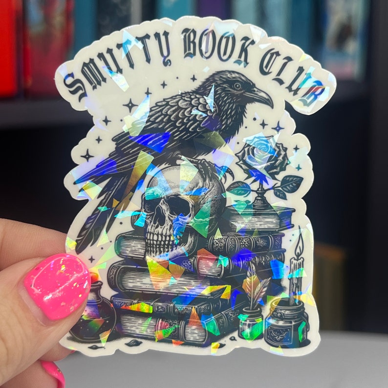 May include: A holographic sticker with a black raven, skull, and stack of books. The sticker says "Smutty Book Club".