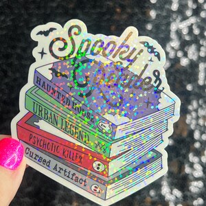 Op de afbeelding: Een holografische sticker met de tekst "Spooky Reader" en een stapel boeken met de titels "Haunted House", "Urban Legend", "Psychotic Killer" en "Cursed Artifact".